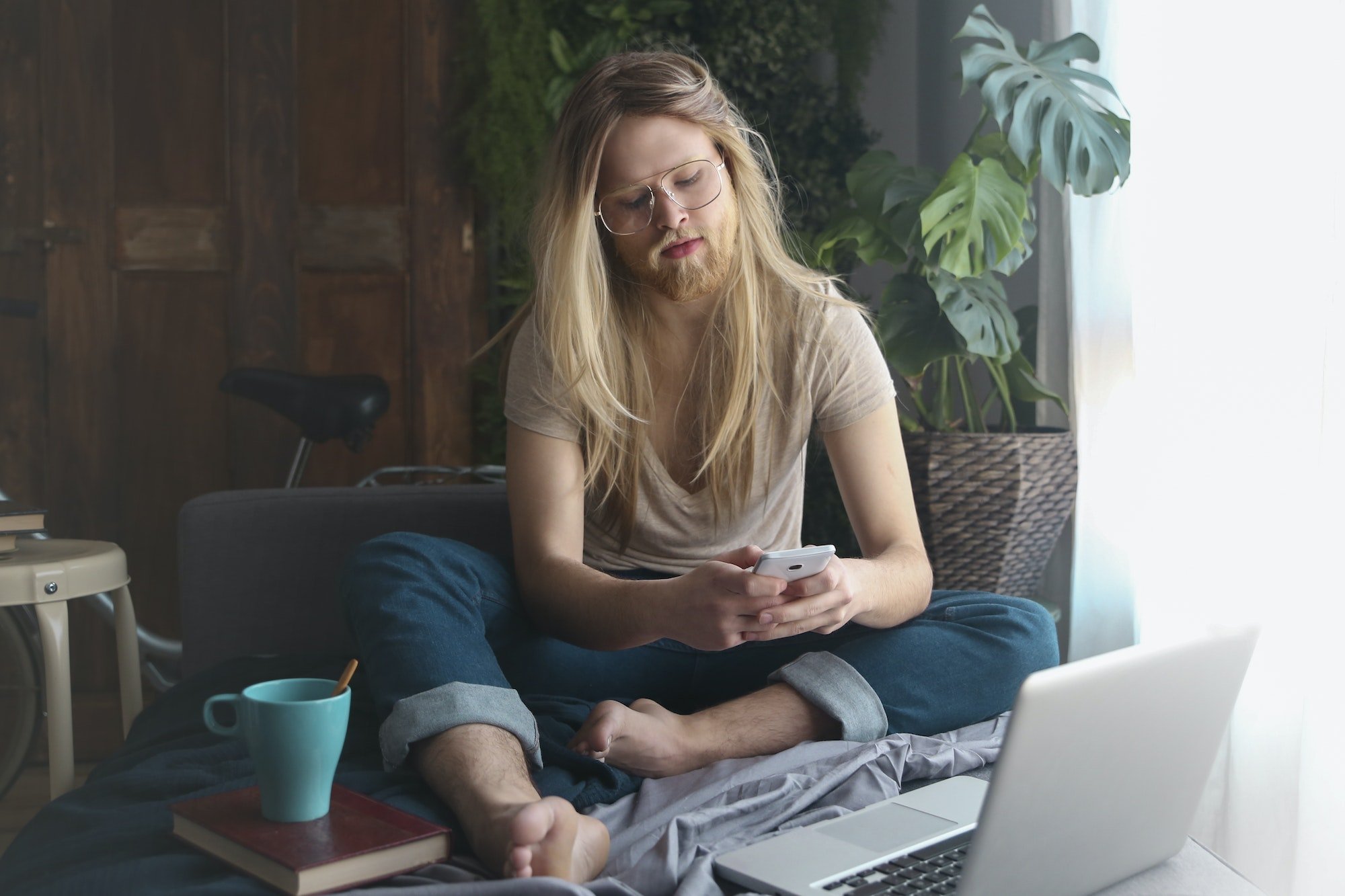 Man with long hair and beard sitting on sofa bed looking at smartphone